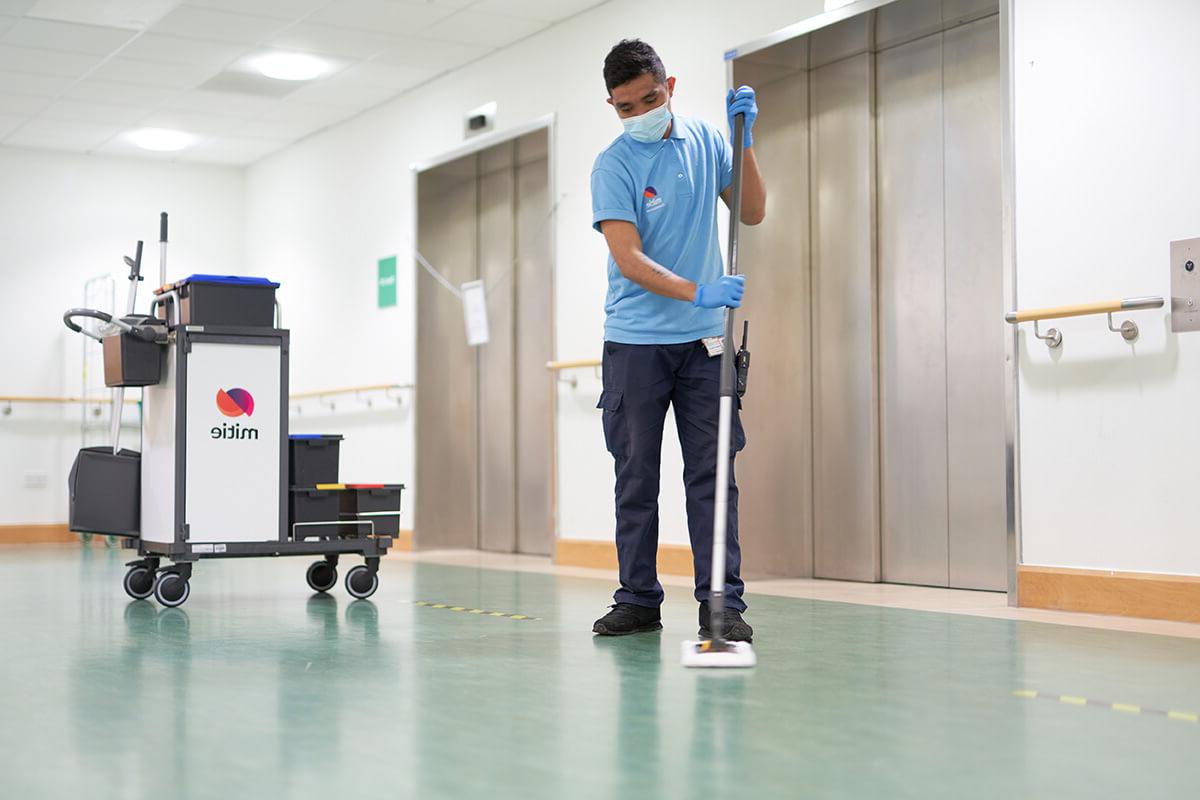 A Mitie employee using a broom to clean the floor in a hospital corridor, with a Mitie branded cleaning cart in the background